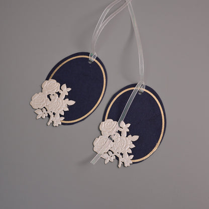 Two navy blue oval fabric tags with gold trim and white floral lace appliqué, hanging from clear ribbons against a plain gray background.