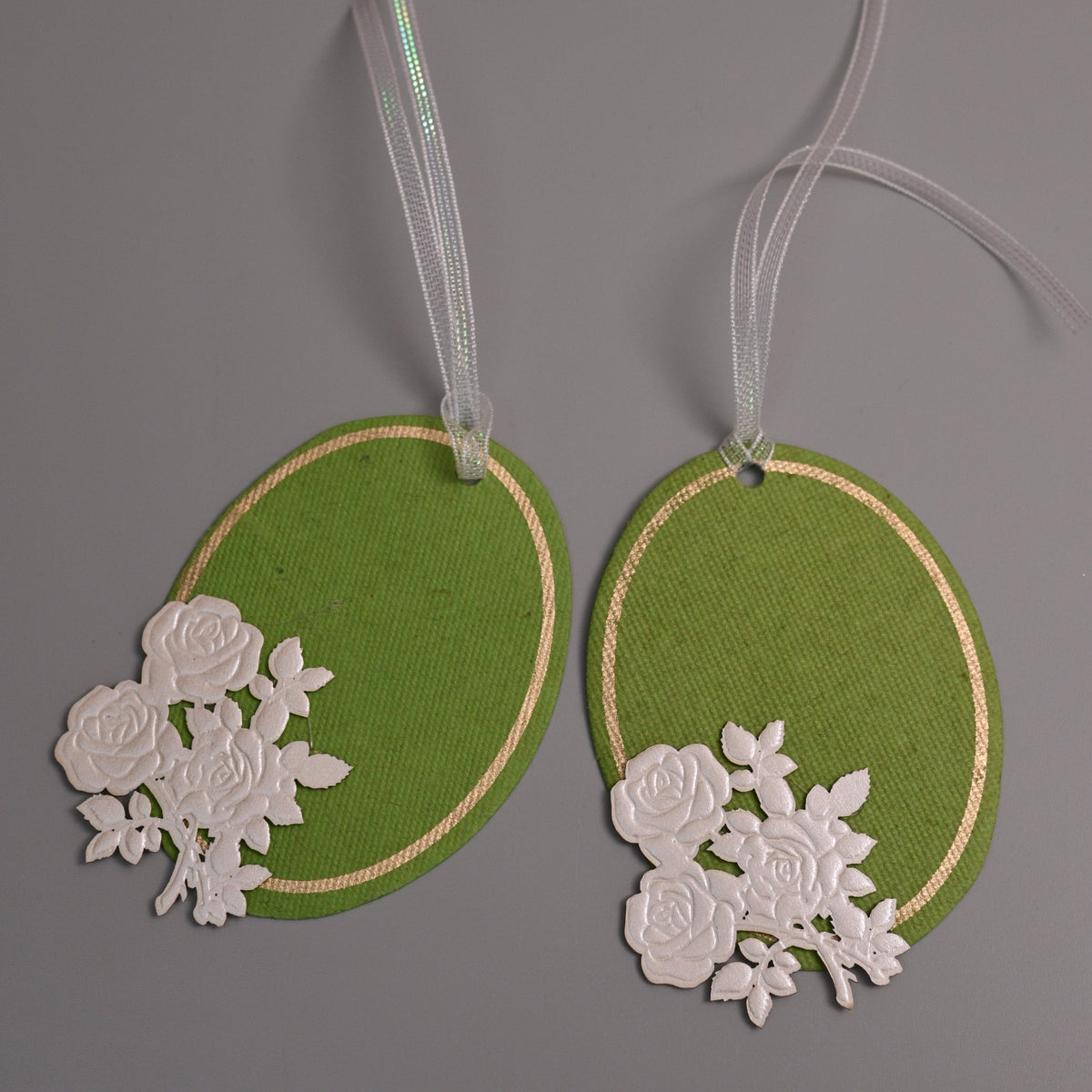 Two green oval gift tags with white floral cutout embellishments and white ribbons