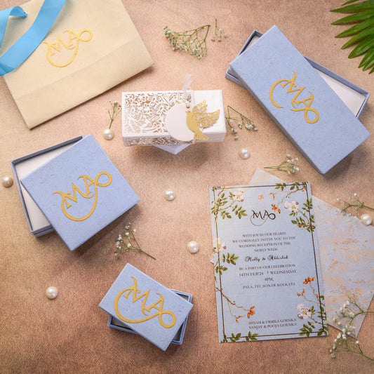 Wedding invitation set with blue boxes, floral design, and gold monogram on a textured surface.