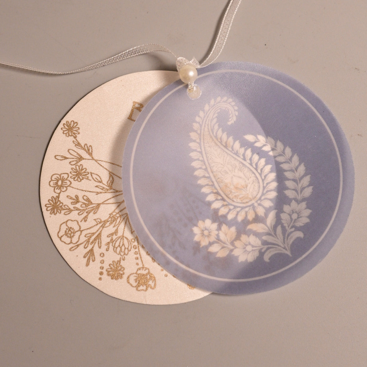 Two round hanging tags on a thin white string, one in translucent lavender with a white paisley and floral design, and one in cream with a gold floral print.