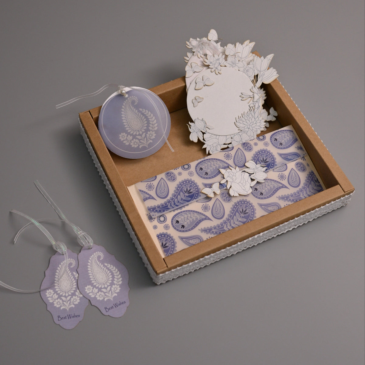 A decorative gift set arranged in a small box on a gray background, including a round lavender ornament with a white paisley design, a white floral cutout, patterned paper pieces, and two matching lavender hanging tags with white ribbons.