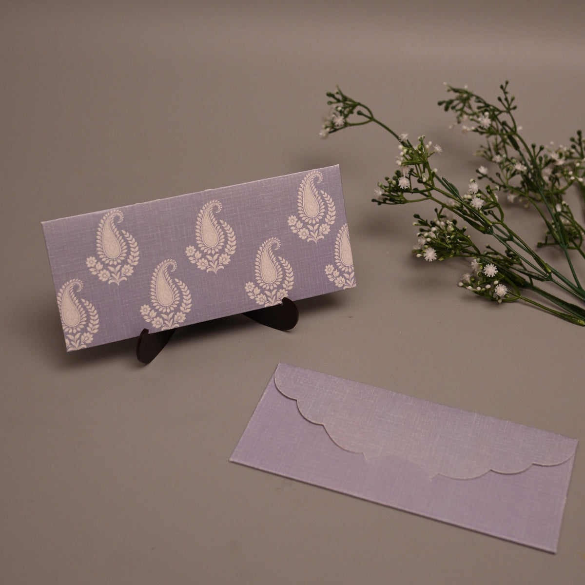 A lavender and white patterned envelope set displayed on a neutral background, featuring one envelope standing upright on a small black stand and another lying flat below it, with small white flowers to the right.