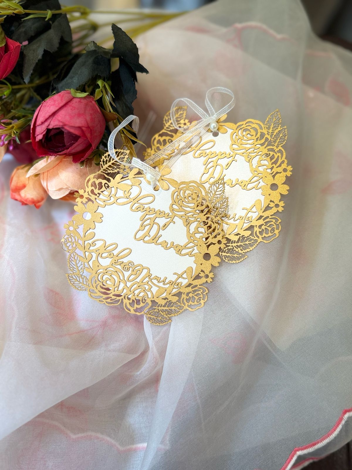 Exquisite laser-cut 'Happy Diwali' gift tags with gold floral and leaf cutouts on a white background, attached with a satin ribbon and a pearl bead.