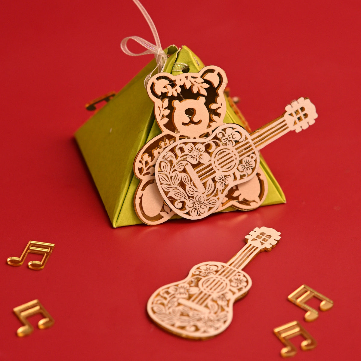 An acrylic bear-shaped guitar ornament with a hanging loop, displayed on top of a green pyramid-shaped box with small gold musical note and guitar-shaped confetti scattered around.