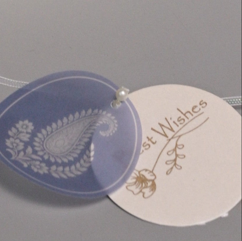 A round translucent lavender-blue tag with a white paisley and floral design, overlapping a cream-colored round tag printed with the words 'Best Wishes' in gold script.