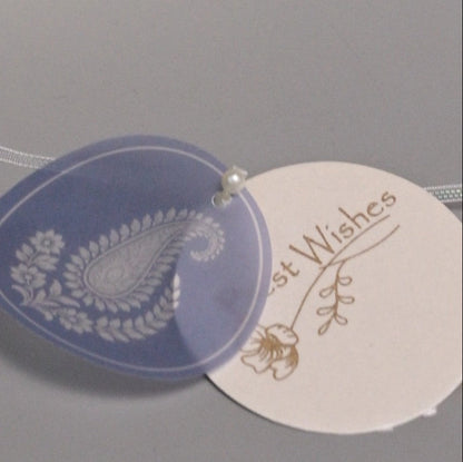 A round translucent lavender-blue tag with a white paisley and floral design, overlapping a cream-colored round tag printed with the words 'Best Wishes' in gold script.