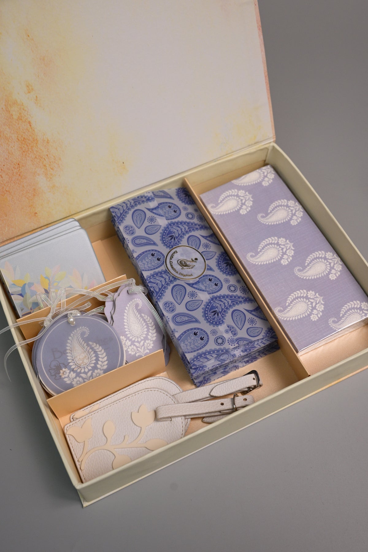 A gift set in a beige box with a watercolor-style printed interior lid, containing a blue paisley-patterned wallet, a lavender paisley-patterned card holder, a matching zippered pouch, a small round tag, and luggage tags
