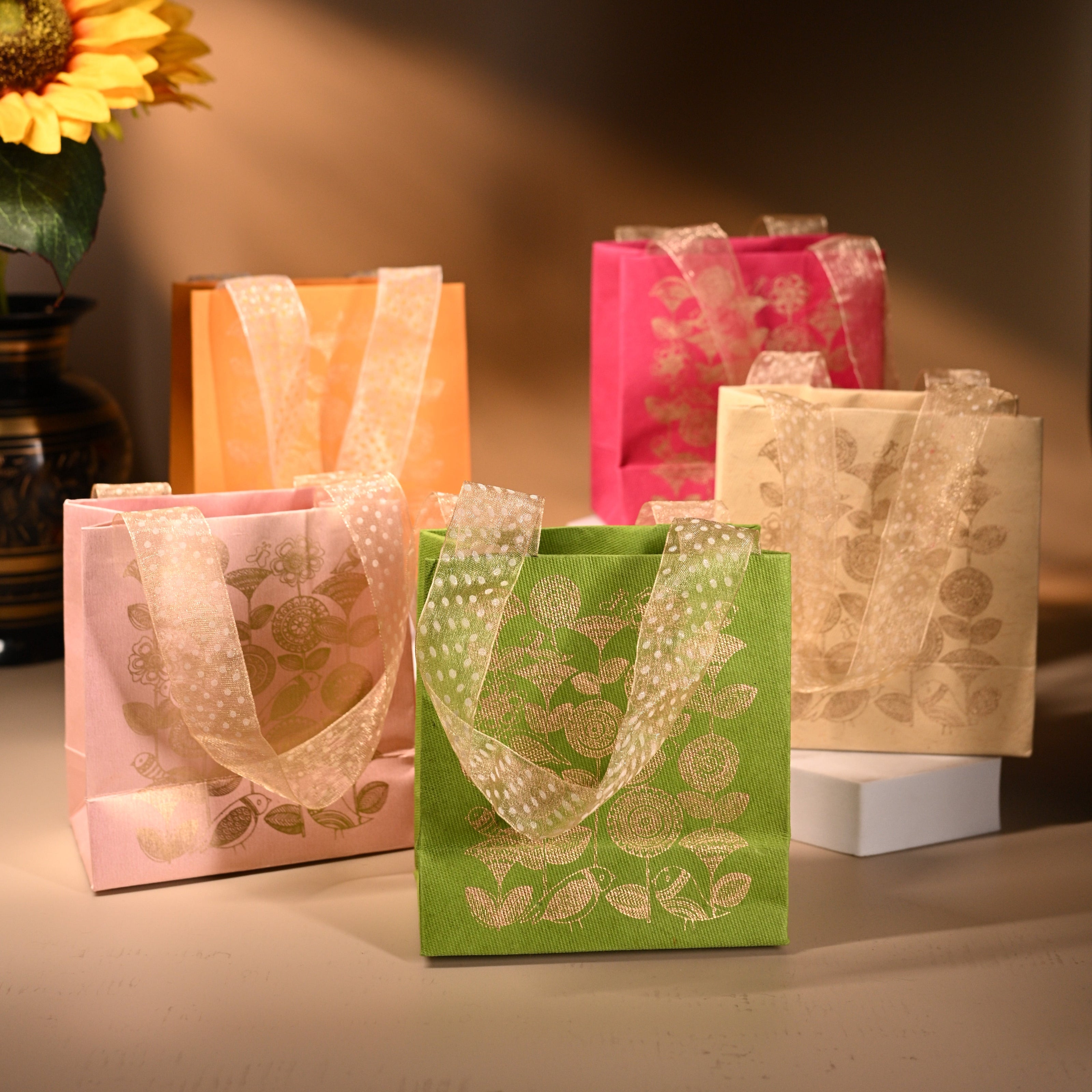 A set of five handmade paper bags in various colors, each featuring a gold floral print and ribbon handles.