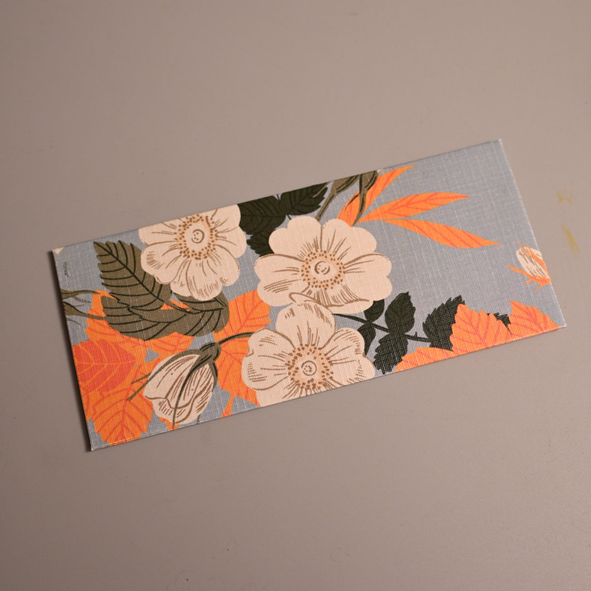 Money Envelope with a floral pattern featuring cream flowers, orange leaves, and dark green foliage on a light blue-gray background, placed diagonally on a plain taupe surface. 