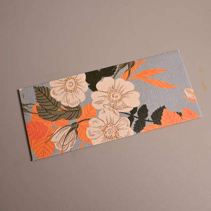Money Envelope with a floral pattern featuring cream flowers, orange leaves, and dark green foliage on a light blue-gray background, placed diagonally on a plain taupe surface. 