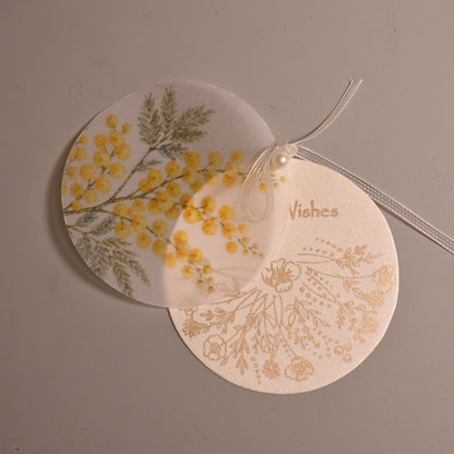 Two round decorative paper tags with botanical designs, one featuring yellow flowers and green leaves on a white background, and the other with gold floral patterns on a cream background, tied together with a clear string on a neutral gray surface.