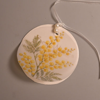 A round butter paper tag with a hand-painted yellow mimosa design, attached to a white cord with a small pearl bead.