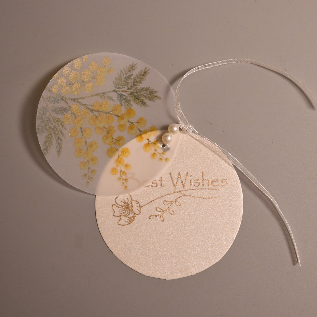 Two layered round gift tags with a thin white ribbon, one printed with yellow flowers and the other with the words 'Best Wishes' in gold foil, on a neutral gray background.