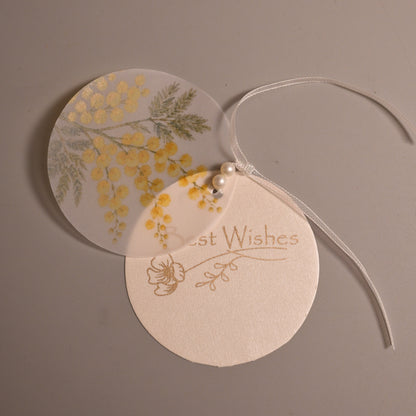 Two layered round gift tags with a thin white ribbon, one printed with yellow flowers and the other with the words 'Best Wishes' in gold foil, on a neutral gray background.