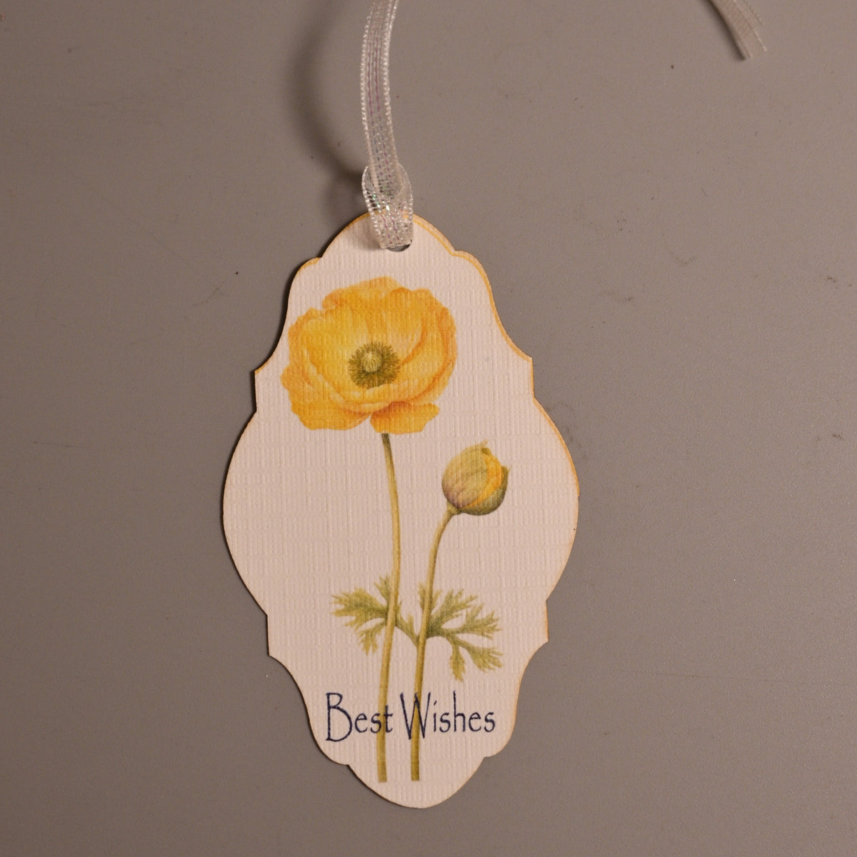 A decorative gift tag with a scalloped edge, featuring a yellow poppy flower illustration and the handwritten text 'Best Wishes' at the bottom.