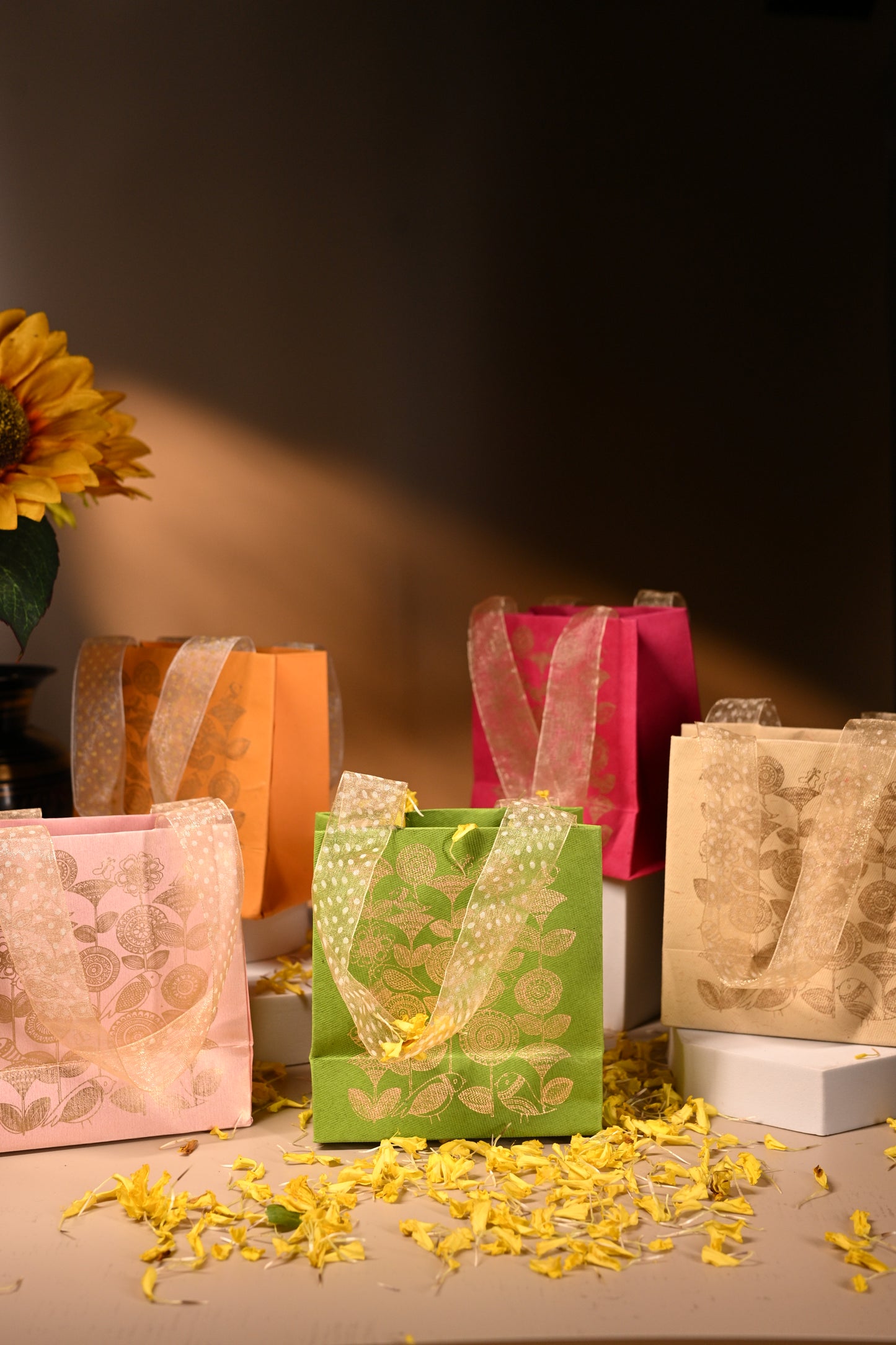 A set of five handmade paper bags in various colors, each featuring a gold floral print and ribbon handles.