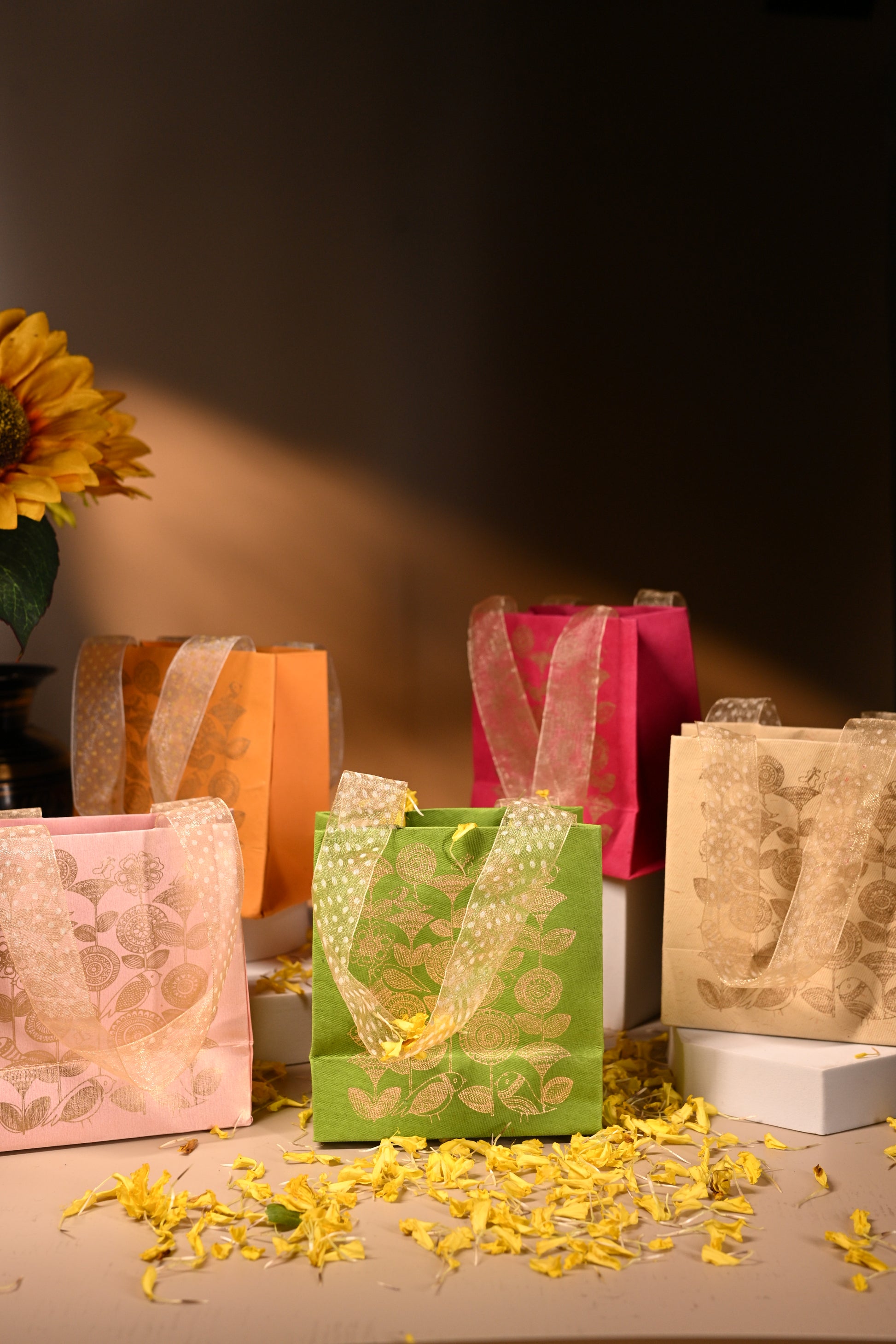 A set of five handmade paper bags in various colors, each featuring a gold floral print and ribbon handles.