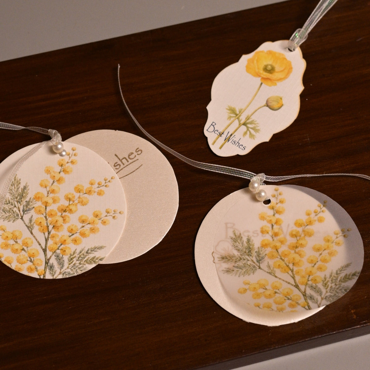 Three round and irregular white paper tags with yellow floral illustrations and thin hanging strings, arranged on a dark brown wooden board against a plain gray background.