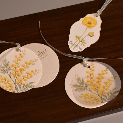 Three round and irregular white paper tags with yellow floral illustrations and thin hanging strings, arranged on a dark brown wooden board against a plain gray background.